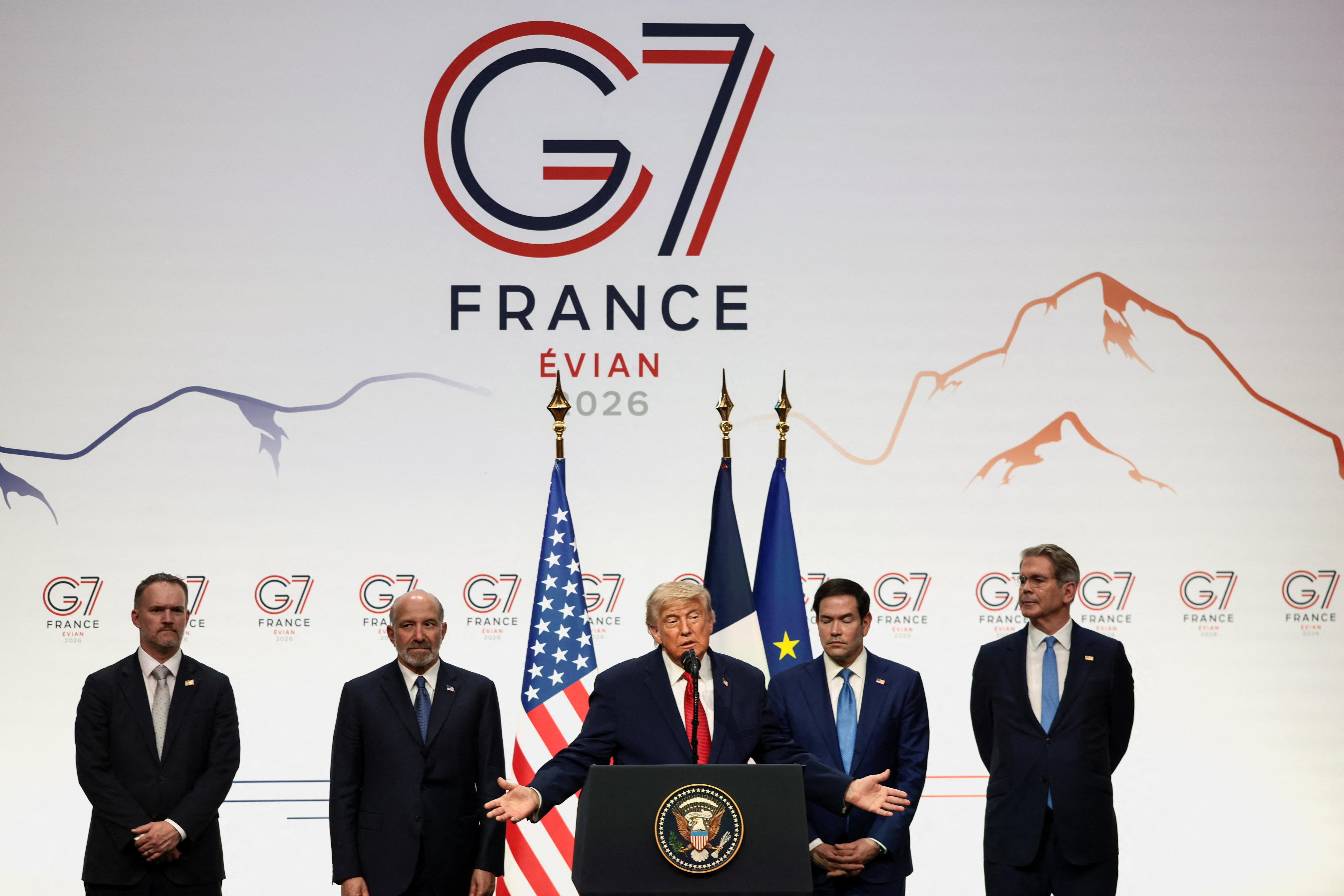 Donald Trump stands at a podium talking and gesturing. Four other men stand on the stage.