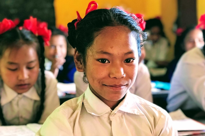 A young Nepalese girl smiles in front of a lot of other students in a classroom. They all have red bows in their hair.