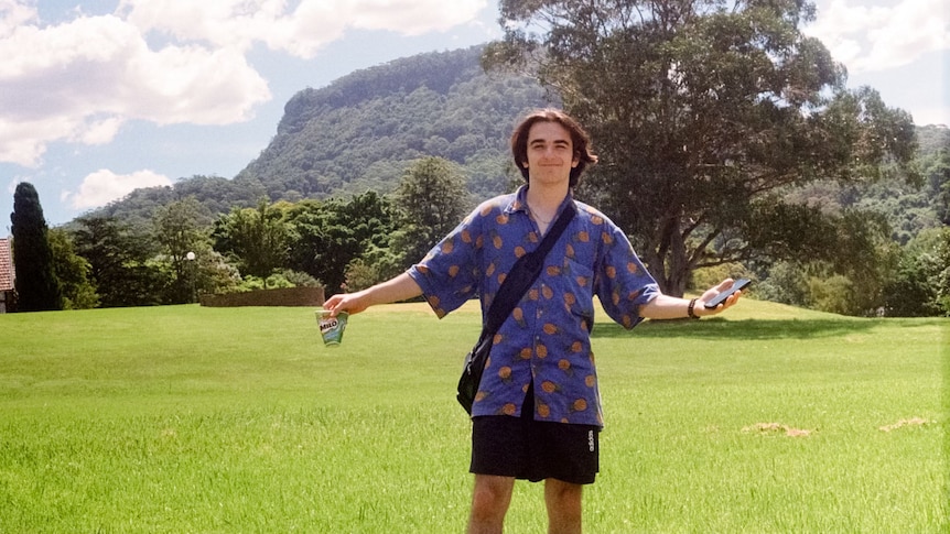 PJ stands in a green field wearing a blue button up and black shoulder bag & shorts.