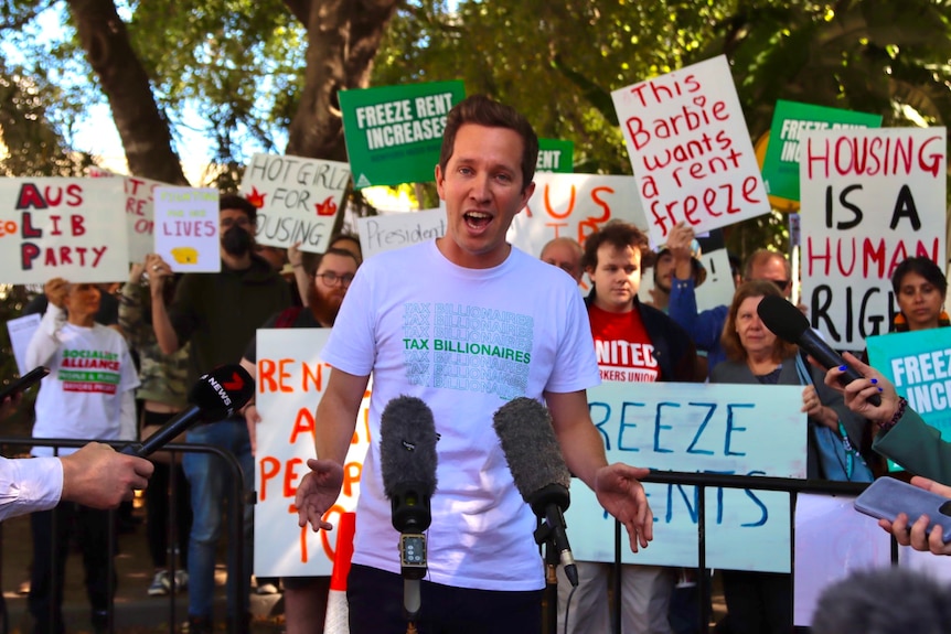 a man speaking at a mic in front of protestors holding signs