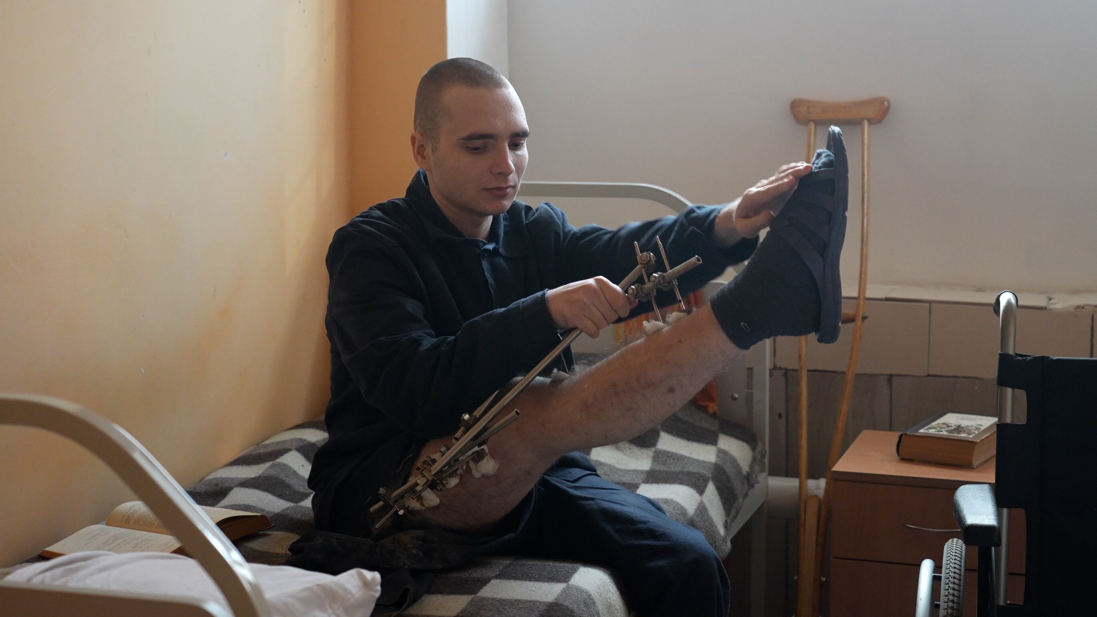 A young man with a shaved head holds up his leg which is in a wire brace.