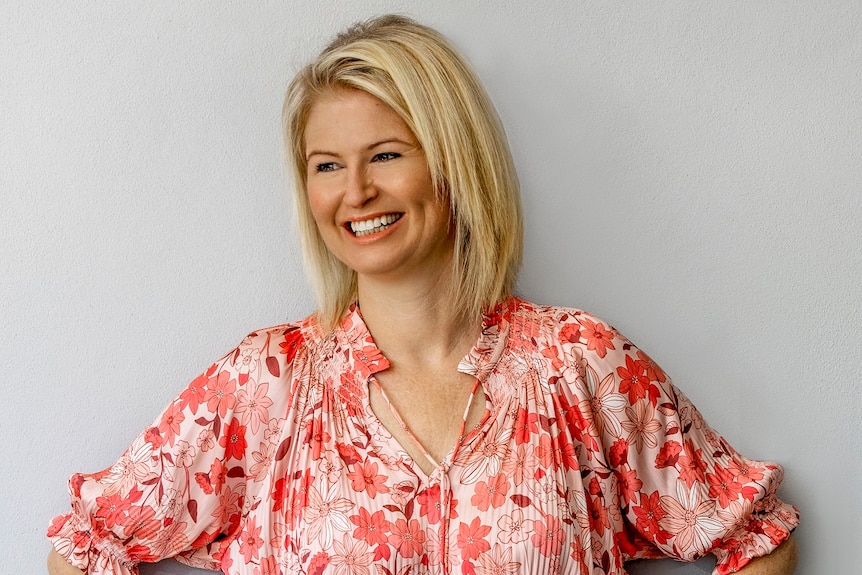 A woman with blonde short hair standing against a wall smiling wearing a pink blouse and white shorts.