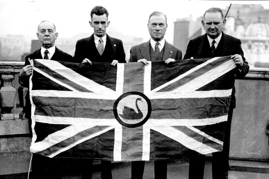 Black and white pic of four men holding the proposed new WA flag - a Union Jack with a black swan in the centre.