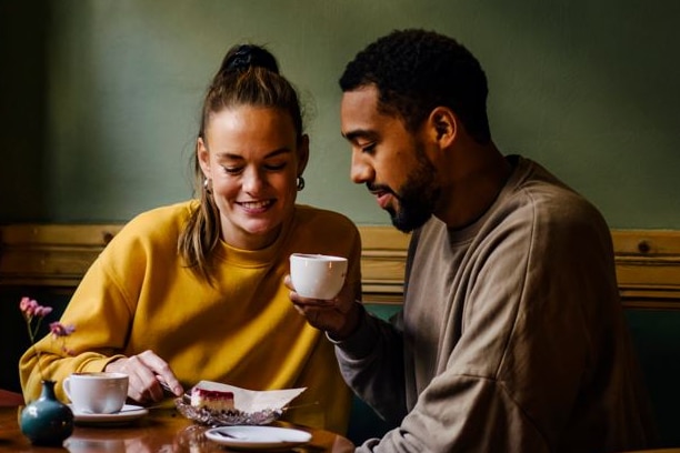 A man and woman sit in dimly lit room with mugs in front of them, both smiling and looking down.