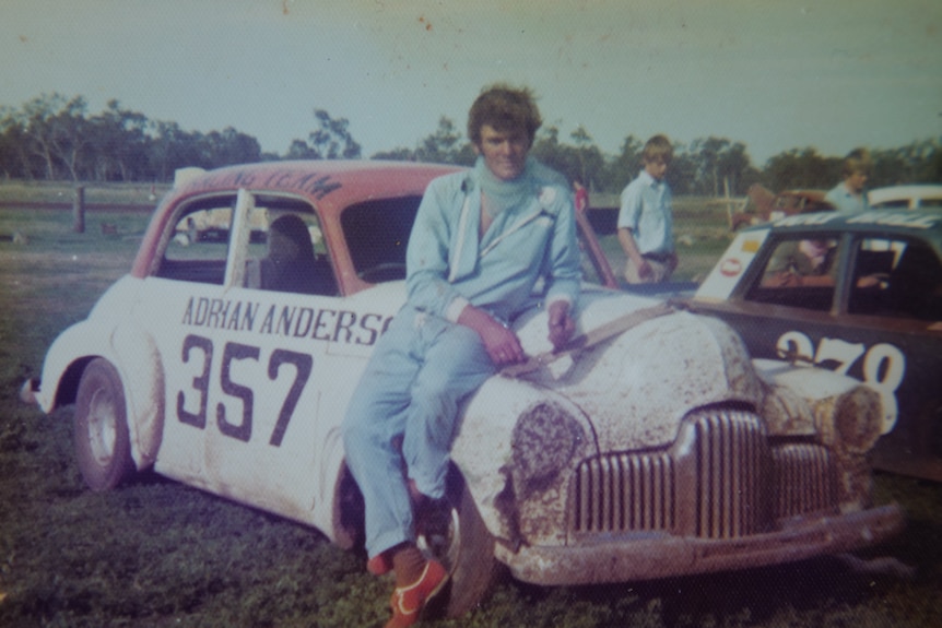 A man in a racing suit leans against the bonnet of a racing car 