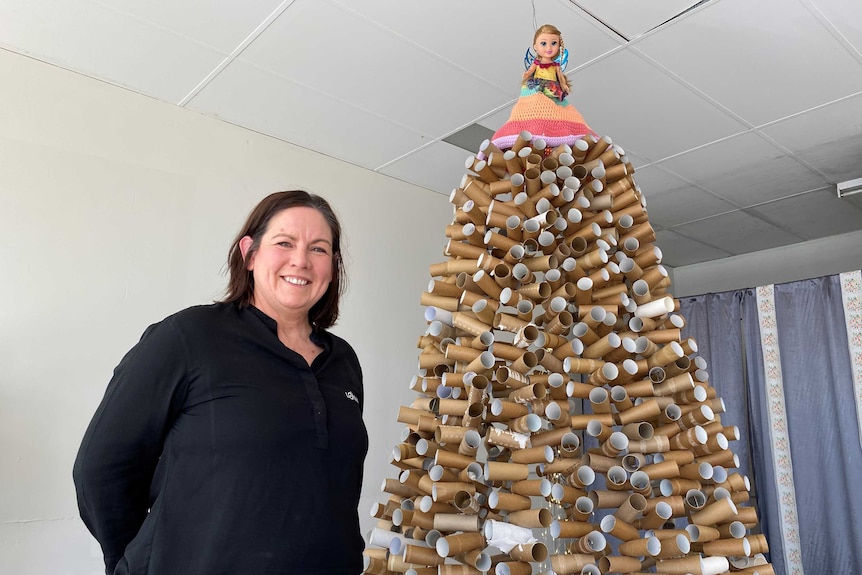 A middle aged woman stands next to a large christmas tree made out of toilet paper rolls