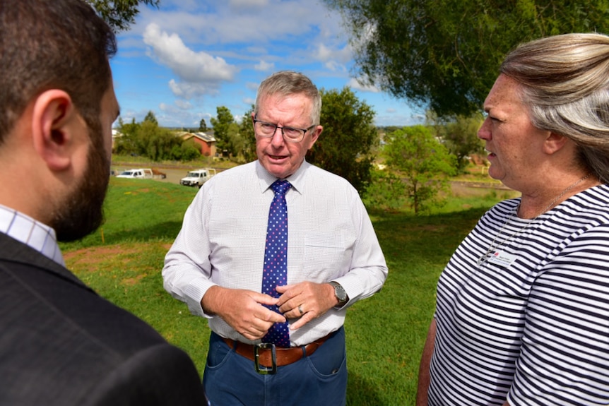 A man wearing a white shirt, blue tie and glasses stands on a patch of grass talking to two people.