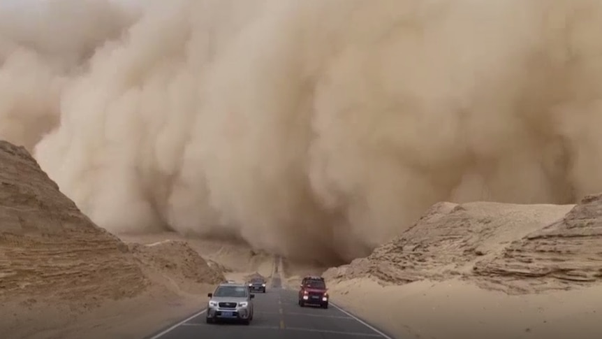 Three cars speed down a desert road away from a gigantic desert sandstorm.