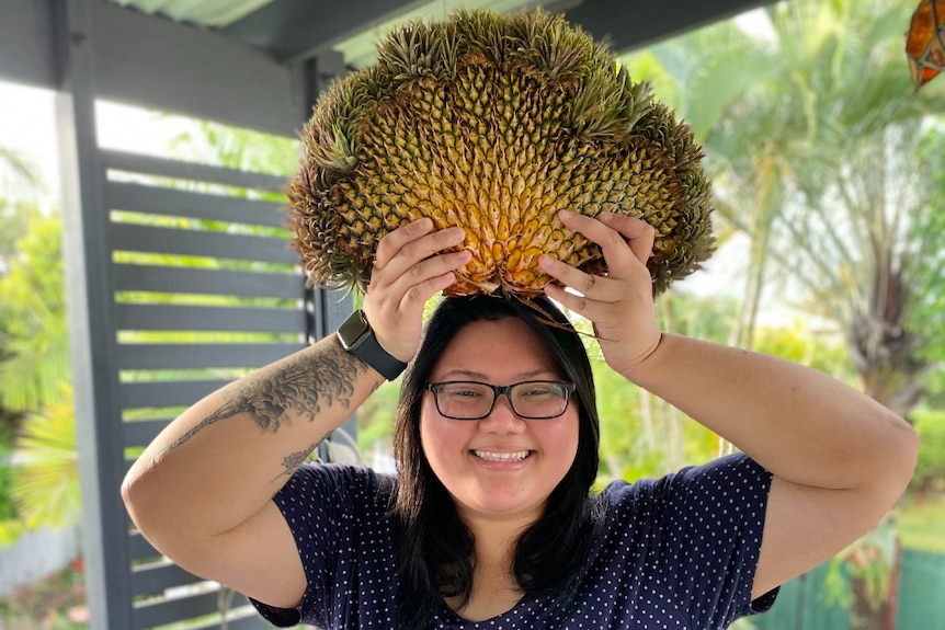 A woman standing with a fasciated pineapple which looks like a fan, balancing on top of her head. She is smiling