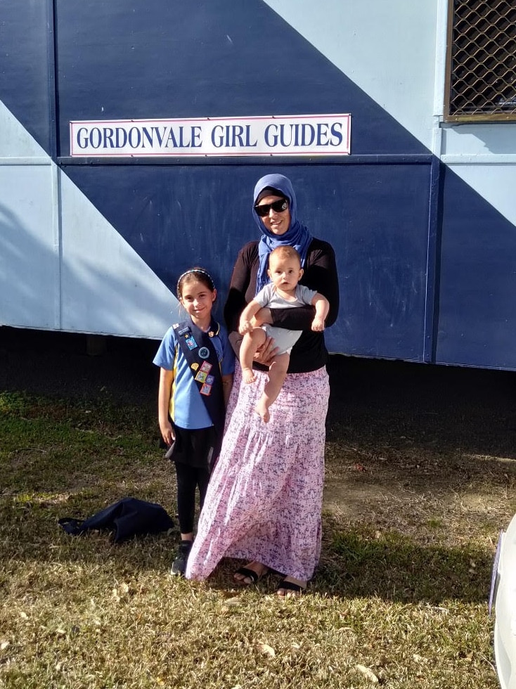 Alma Mohammed stands outside a Girl Guides building. She is holding a baby. Beside her is a young girl in a Girl Guides uniform.