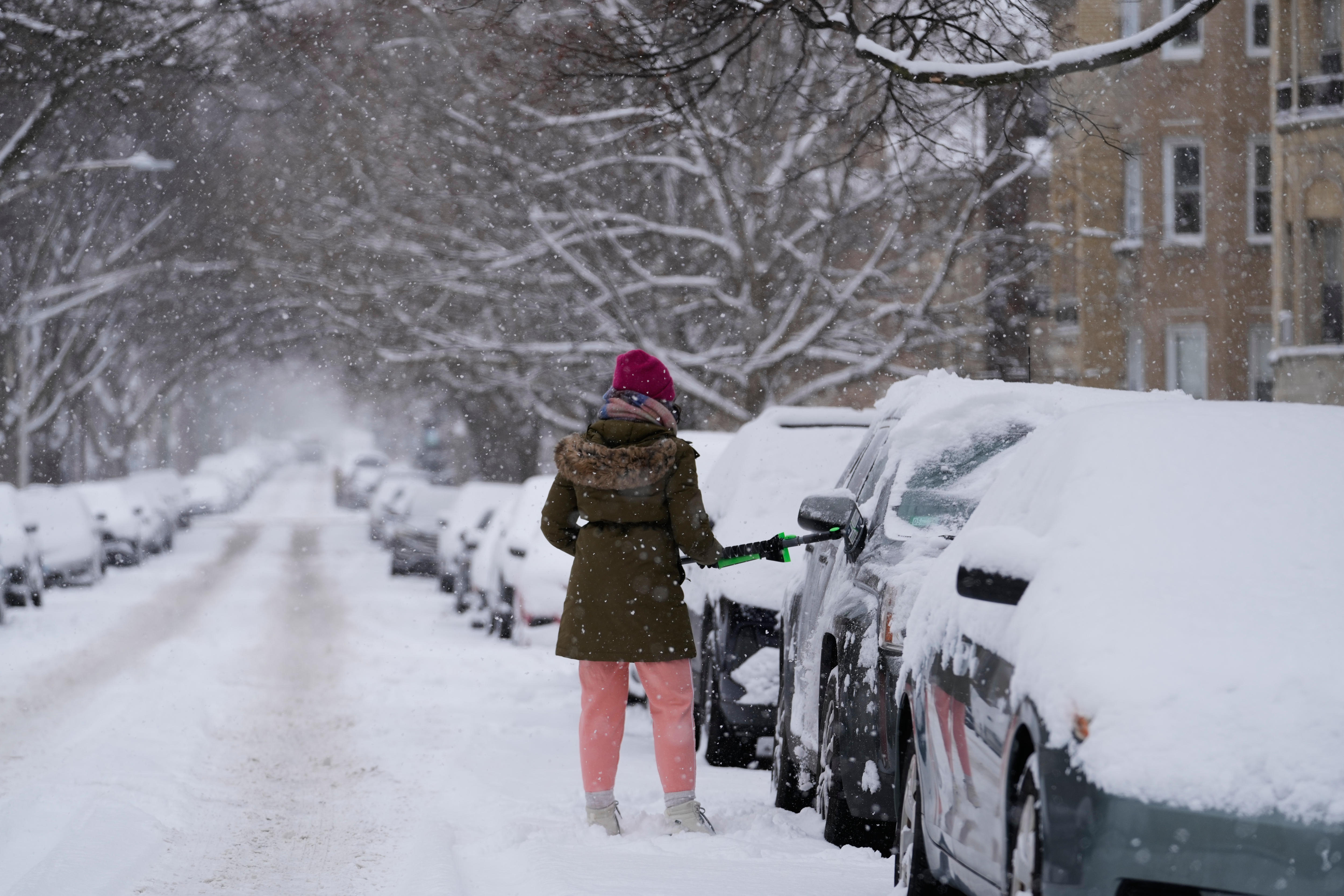 Una mujer quita la nieve de un coche en una calle llena de vehículos cubiertos de nieve.