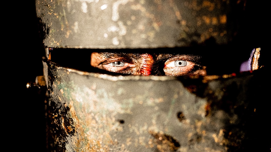 Blue eyes on a bloody face look through a small rectangular slit on large metal facial armour.