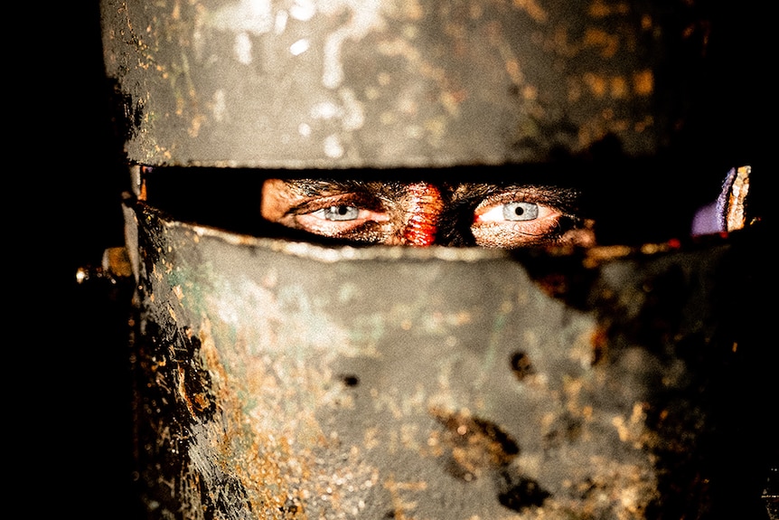 Blue eyes on a bloody face look through a small rectangular slit on large metal facial armour.