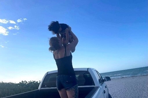 A woman stands on a beach holding her daughter up in the air in an embrace.