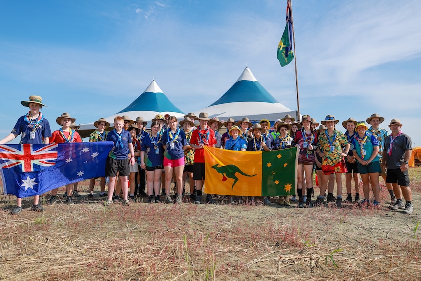 A large group of young people in scout uniforms, holding up Australian and boxing kangaroo flags in front of conical marquees.