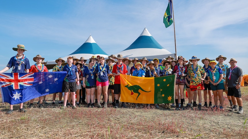 A large group of young people in scout uniforms, holding up Australian and boxing kangaroo flags in front of conical marquees.