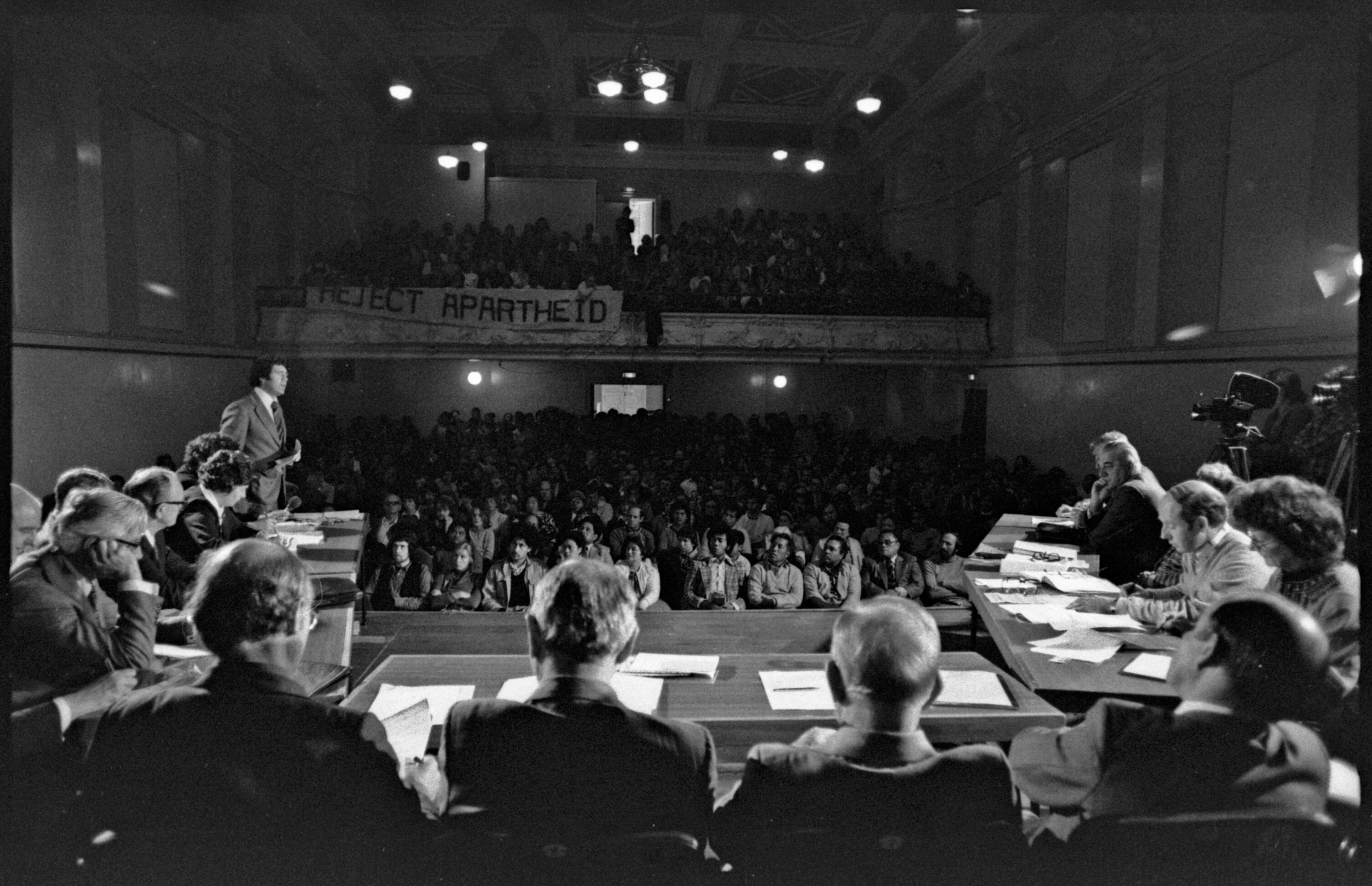 A black and white image shows a meeting on stage with a packed audience. 