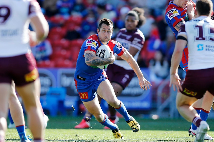 A Newcastle Knights NRL player holds the ball with his right arm tucked into his chest as he runs towards Manly opponents.