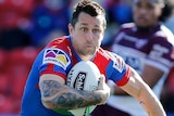 A Newcastle Knights NRL player holds the ball with his right arm tucked into his chest as he runs towards his Manly opponents.