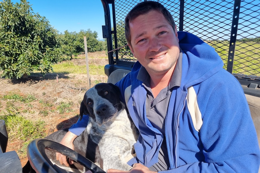 Plan moyen d'un homme dans un VTT tenant son chien devant des avocatiers.