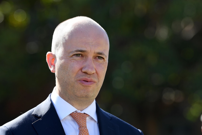 A bald man in a dark suit, orange tie, white shirt talking, background blurred.