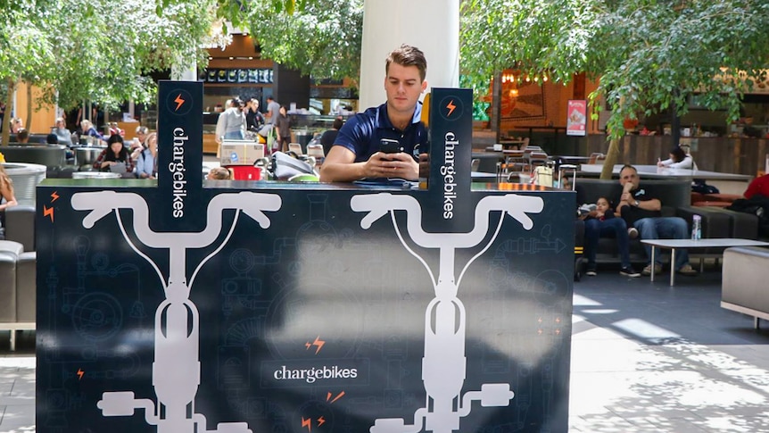 Man sits at a bike kiosk pedalling to charge his mobile phone at Brisbane Airport.