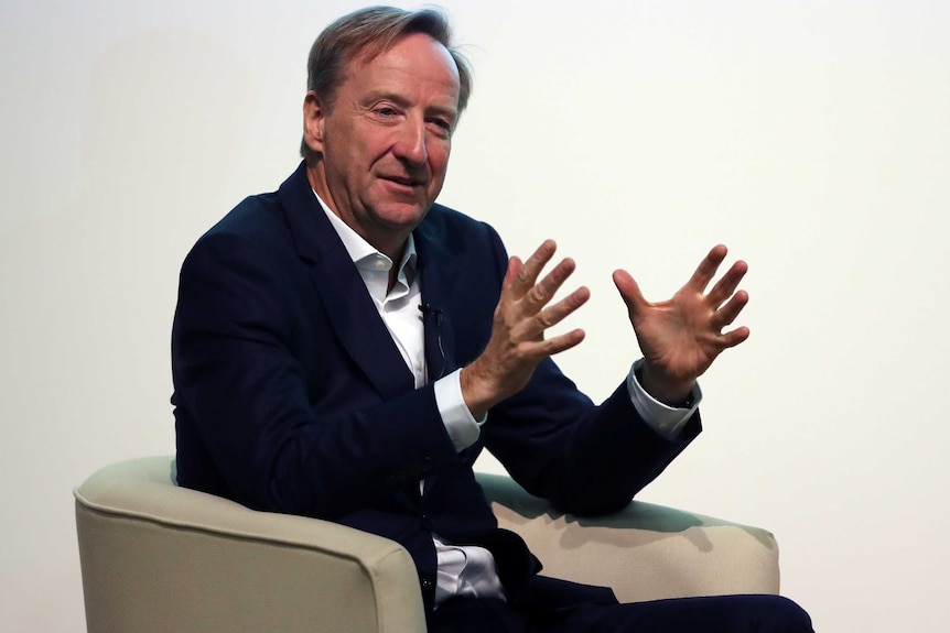 Alex Younger dressed in a suit without a tie sits in a chair.