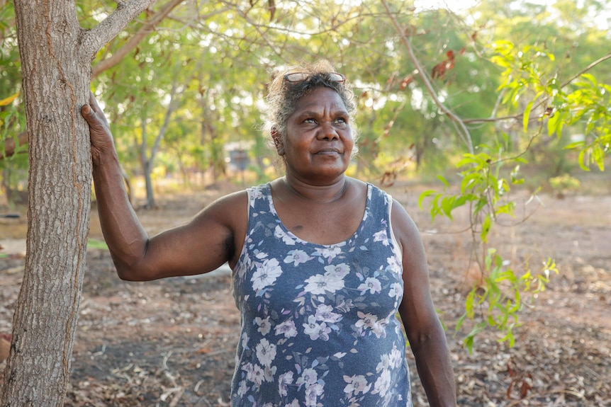 Freda Ali, a Burarra woman in her 50s wearing a navy blue singlet with white flowers on it, stands leaning against a tree trunk