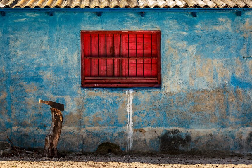 A faded blue concrete wall with a closed, red wooden window in the middle.