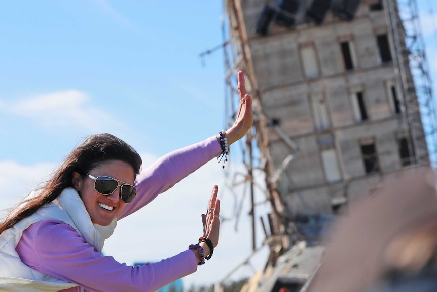 A young woman smiling and wearing sunglass extends her arms in front of a leaning building.