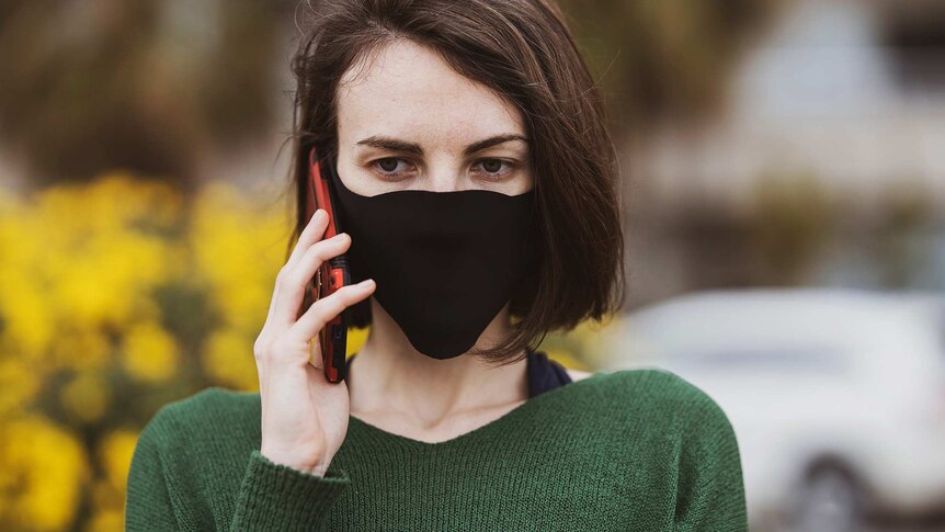 A woman wearing a black mask and green jumper on the phone in a story about what not to say to people in coronavirus lockdown.