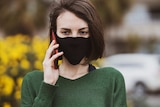 A woman wearing a black mask and green jumper on the phone in a story about what not to say to people in coronavirus lockdown.
