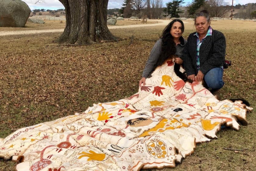 Two indigenous women under a tree displaying a possum skin cloak the size of a single doona