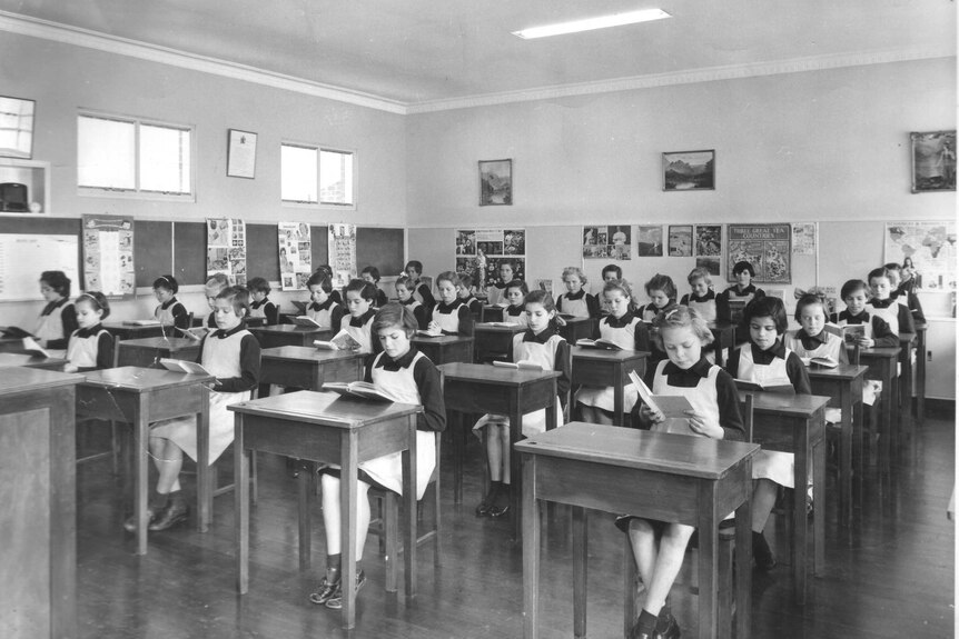 Black and white image of a class of young girls sitting at desks reading books in a classroom