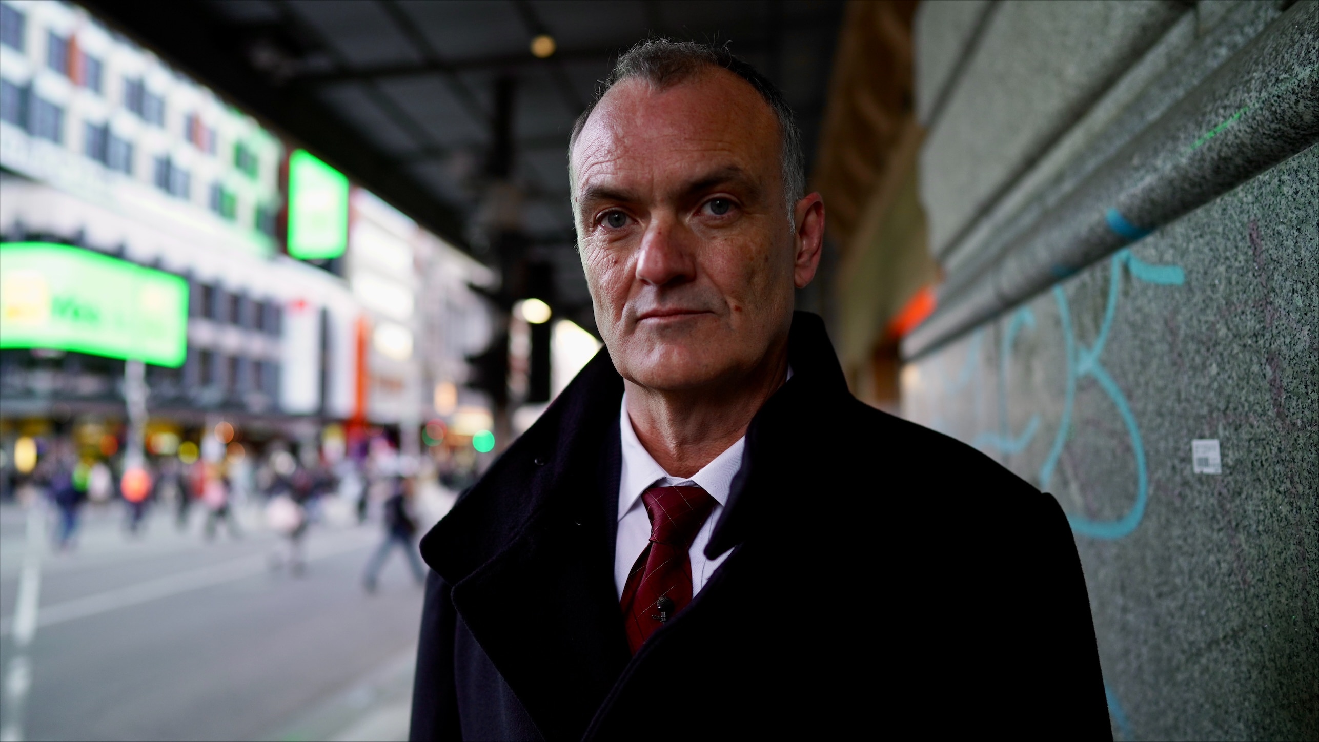 Man wearing a suit and black jacket standing next to a wall. 