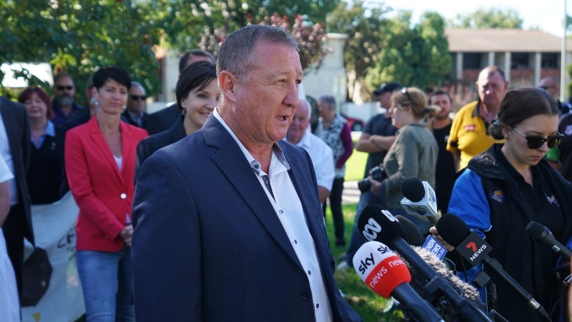 A man in a blue suit stands in front of microphones and a group of people.