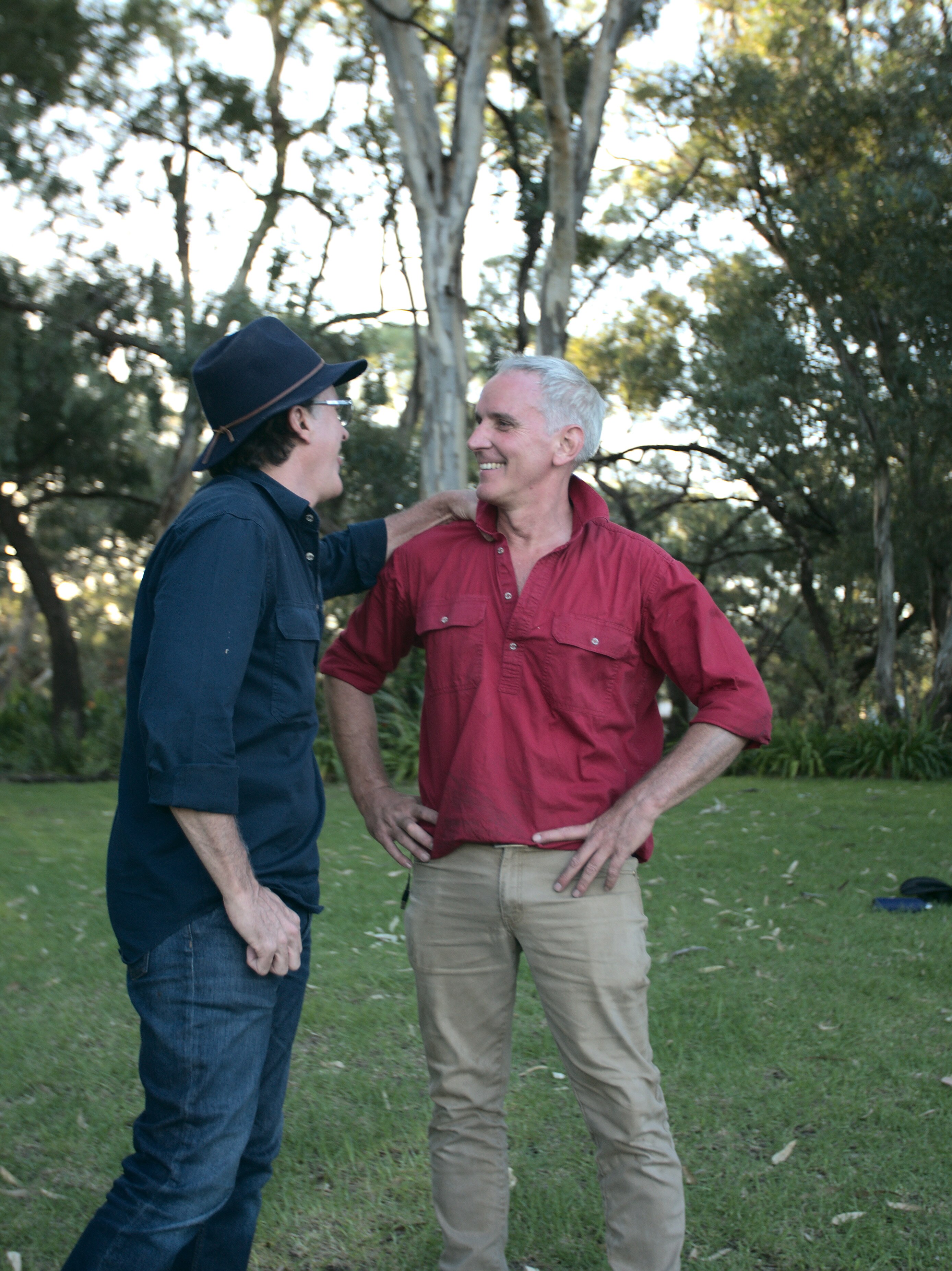 Two men stand in a grassy garden, laughing together.