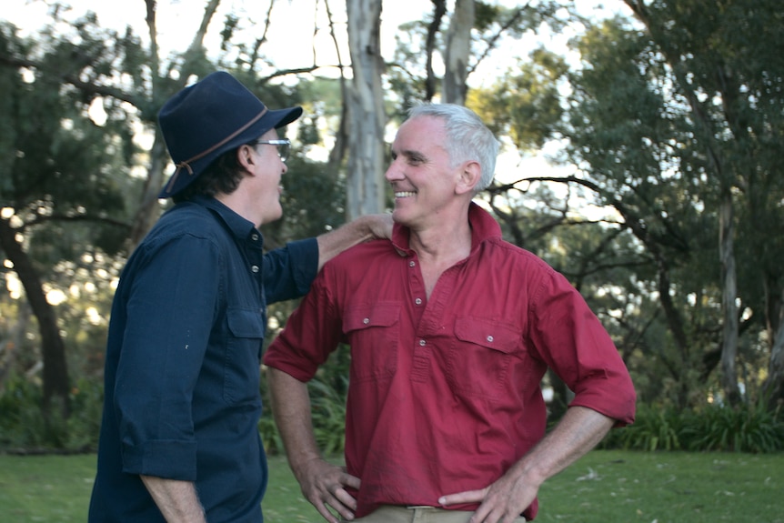 Deux hommes se tiennent dans un jardin herbeux, riant ensemble.