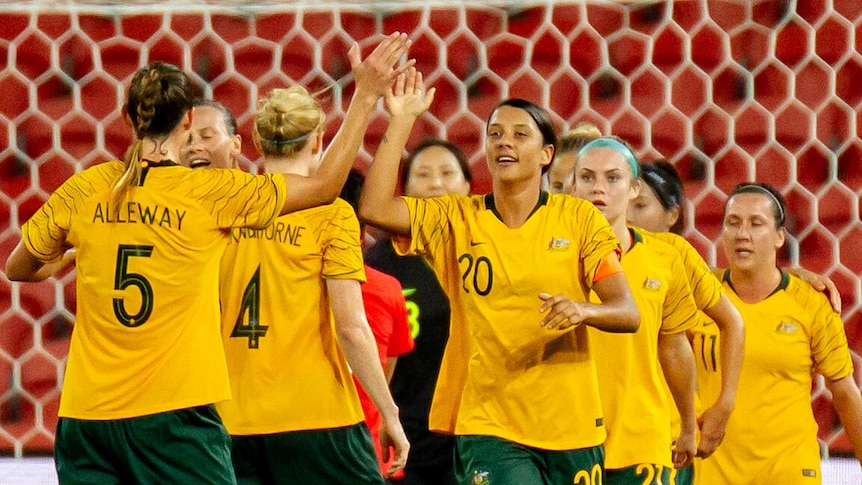 Sam Kerr and Laura Alleway high five with teammates and the goal net in the background.