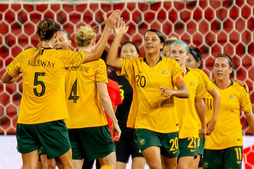 Sam Kerr and Laura Alleway high five with teammates and the goal net in the background.