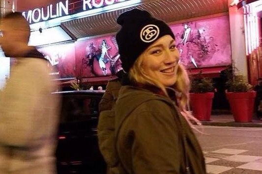 Madison Lyden poses in front of the Moulin Rouge, in a warm coat and beanie, at night, smiling at the camera