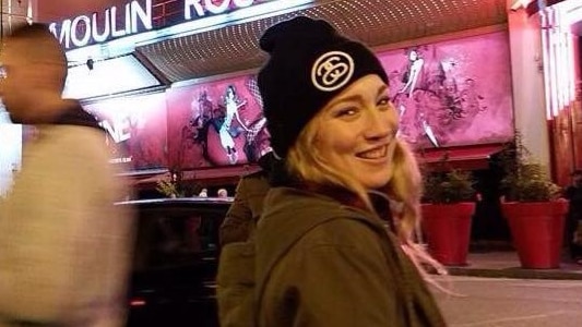 Madison Lyden poses in front of the Moulin Rouge, in a warm coat and beanie, at night, smiling at the camera