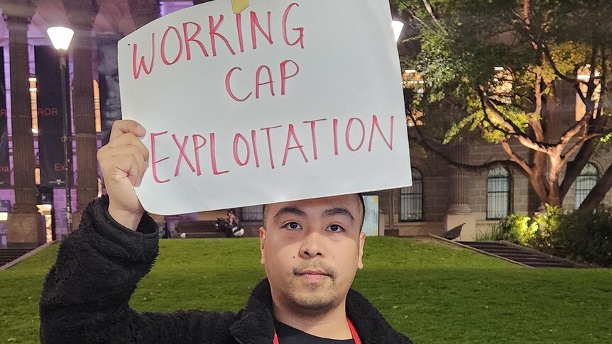 A man holds a sign above this head that says "working cap exploitation" 