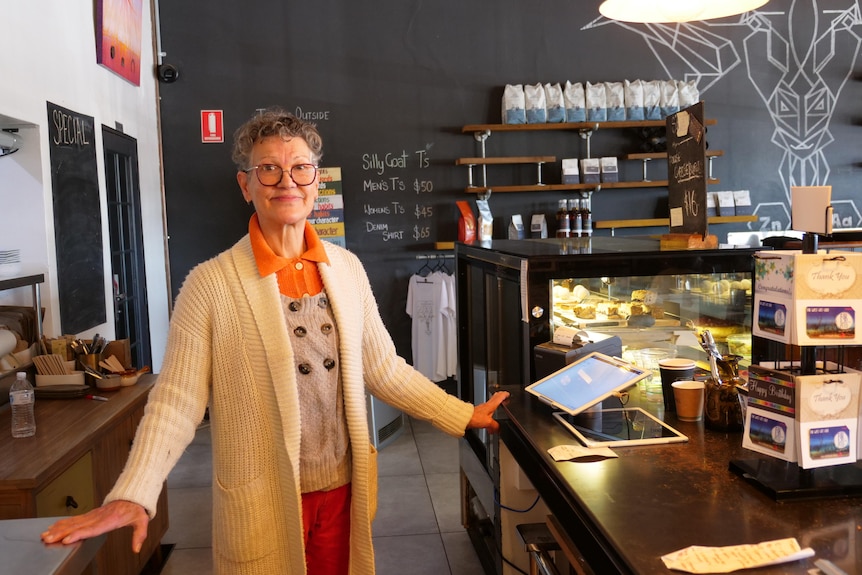 A woman with short, grey hair, wearing bright clothes, stands behind a cafe counter.
