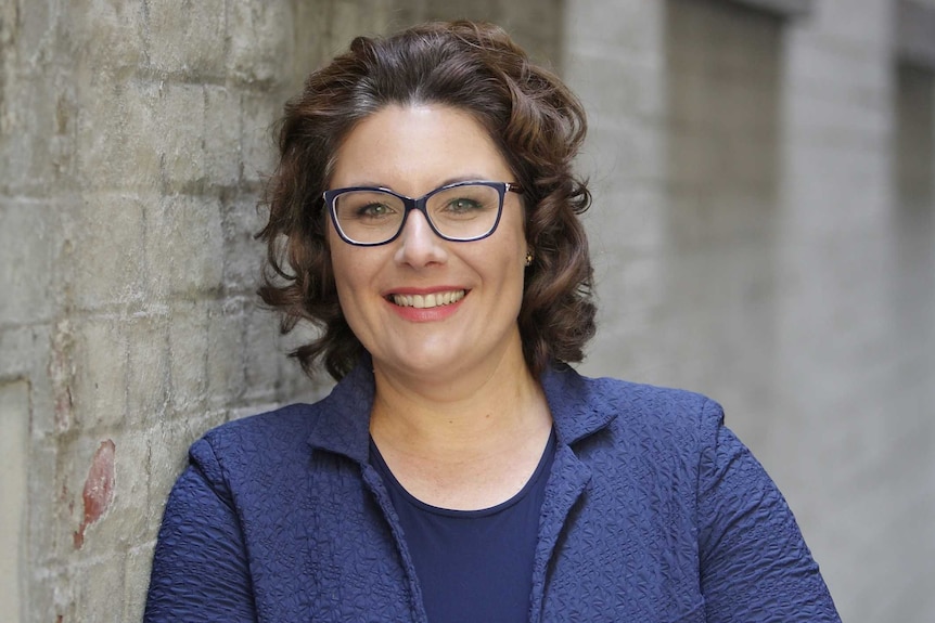 A woman with curly brown hair, glasses and blue jacket leans against a brick wall smiling widely.