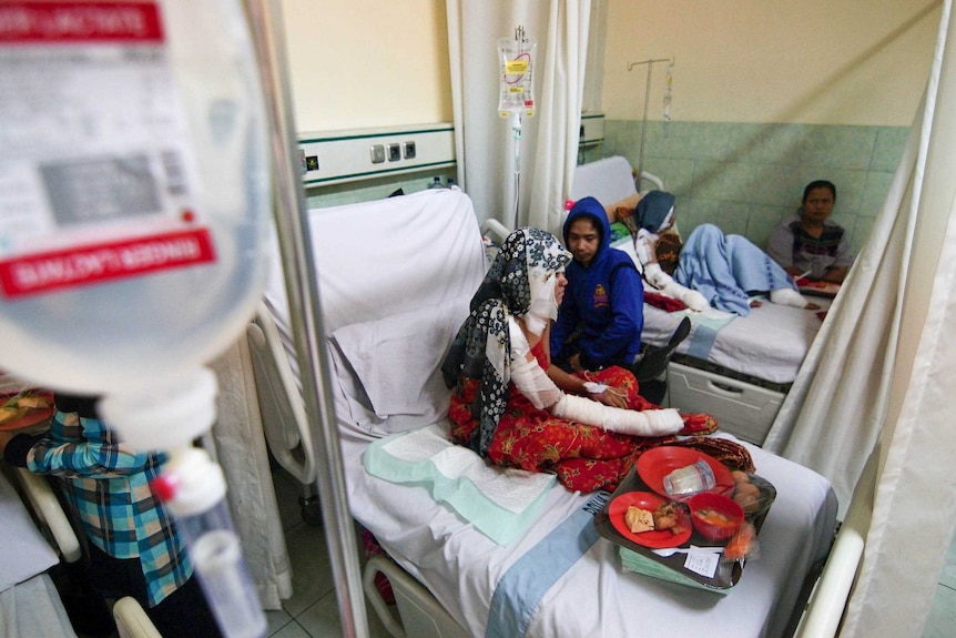 A young woman sits on a hospital bed with a tray of food in front of her, her husband sits beside her
