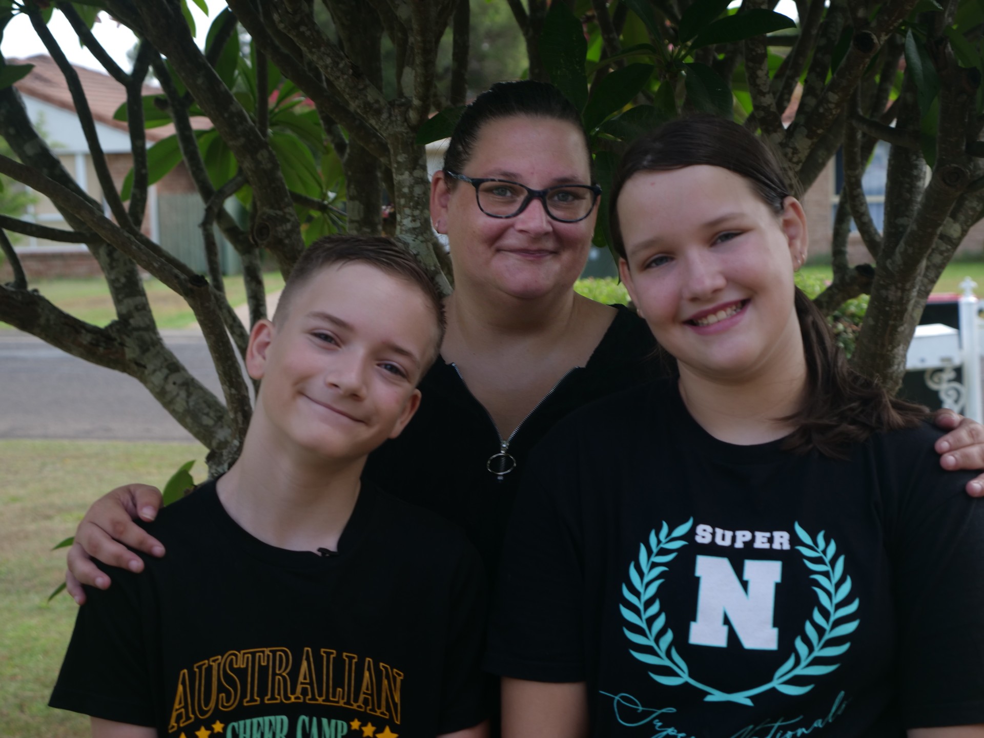 Mother and two teenage children smiling in front of a tree.