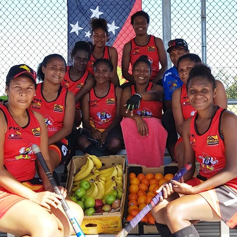 A group of women sit in a grandstand in hockey uniforms around boxes of fruit.