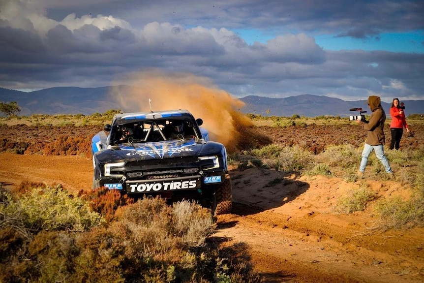 A man films a rally car kicks up a cloud dust as it drifts down a dirt track 