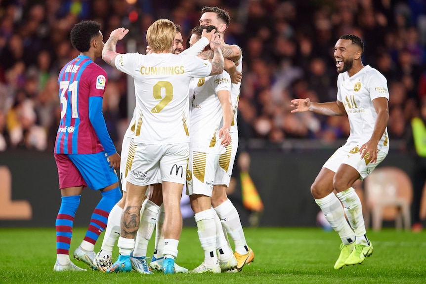A League players in white huddle in celebration. One Barcelona player walks behind.